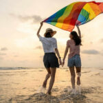 Two people with rainbow flag on beach at sunset.