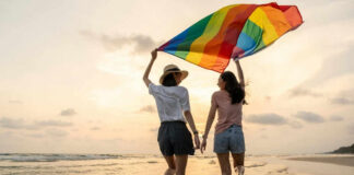 Two people with rainbow flag on beach at sunset.