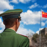 Top Brass Vanish As Xi Tightens Grip Soldier in a green military uniform facing the Great Wall of China with the Chinese flag in the background