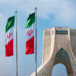 Iran’s New Leader VANISHES — Leg Lost? Coma? Three Iranian flags in front of the Azadi Tower against a blue sky
