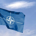 Blue NATO flag with white emblem under cloudy sky.