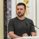 Man speaking at podium with microphones, Ukrainian flag behind.