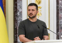 Man speaking at podium with microphones, Ukrainian flag behind.