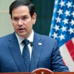 Marco Rubio SLAMS China’s Maritime ‘Bullying’ A man in a blue suit speaking into a microphone with an American flag in the background