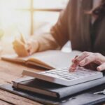 Person calculating taxes with a calculator and writing notes in a notebook at a desk