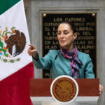 Person speaking at podium with Mexican flag nearby