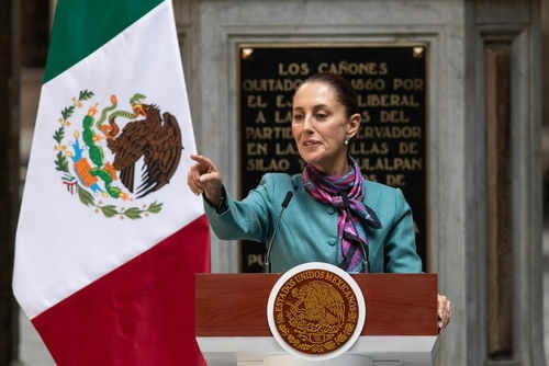 Person speaking at podium with Mexican flag nearby