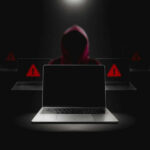 Hacker in hoodie behind laptops with warning signs