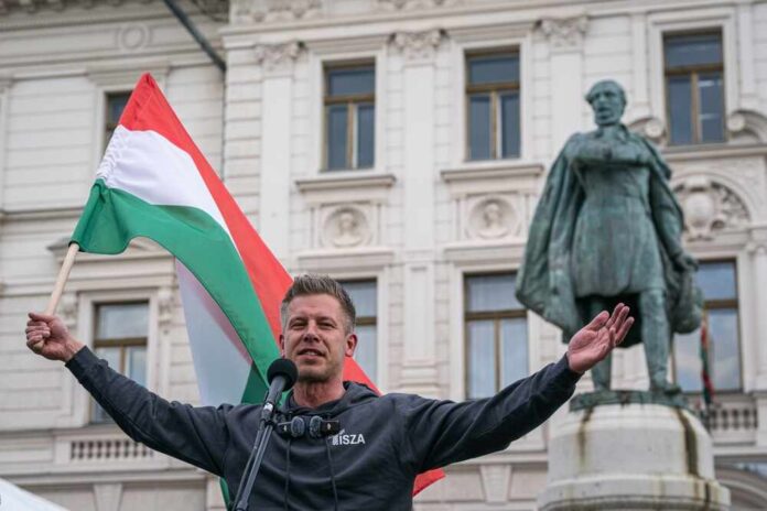 shutterstock_2611175293.jpg Man holding a Hungarian flag while speaking at a rally in front of a statue
