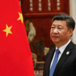 Man in suit standing beside Chinese flag indoors.