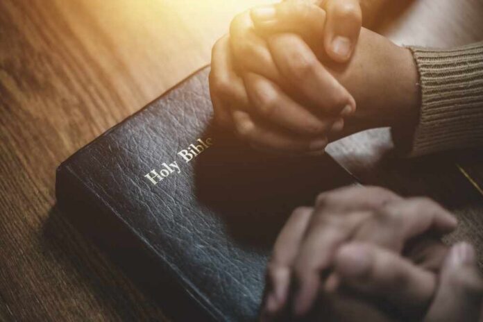 Two hands clasped in prayer over a Holy Bible