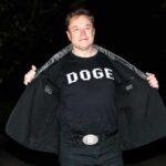 Person wearing a black shirt with 'Doge' printed on it, standing outdoors at night