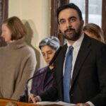 Man in suit speaking at a podium with people standing behind