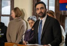 Man in suit speaking at a podium with people standing behind