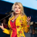 America Pauses: Dolly’s Star Buried In Blooms Singer performing on stage in a bright yellow floral jacket