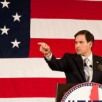 Rubio Drops Hammer On ICC Boss Speaker in suit pointing during speech before large American flag
