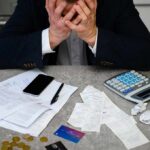 Wallet Squeeze Deepens – More Pain Coming? Stressed person with head in hands at a desk with bills and calculator