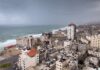 Sleepless Gaza Under Swarming Drones Coastal city skyline with mid-rise buildings by a rough sea under overcast skies
