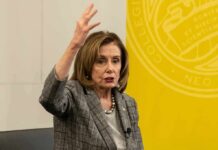 Nancy Pelosi speaking with raised hand in front of a yellow backdrop