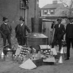 Four men stand around a large metal still with jars and funnels