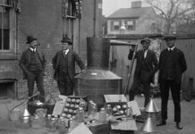 Home Distilling On The Chopping Block Four men stand around a large metal still with jars and funnels