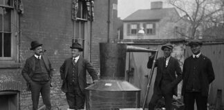 Four men stand around a large metal still with jars and funnels